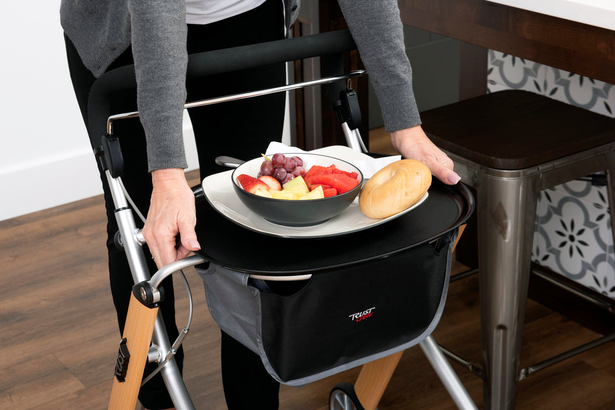 Indoor rollator with attached tray holding a bowl of fruit and bread roll, used by a person indoors near a kitchen counter