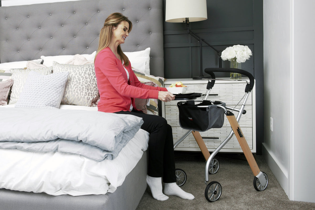 Indoor rollator with tray and storage bag beside a woman seated on a bed in a bedroom setting