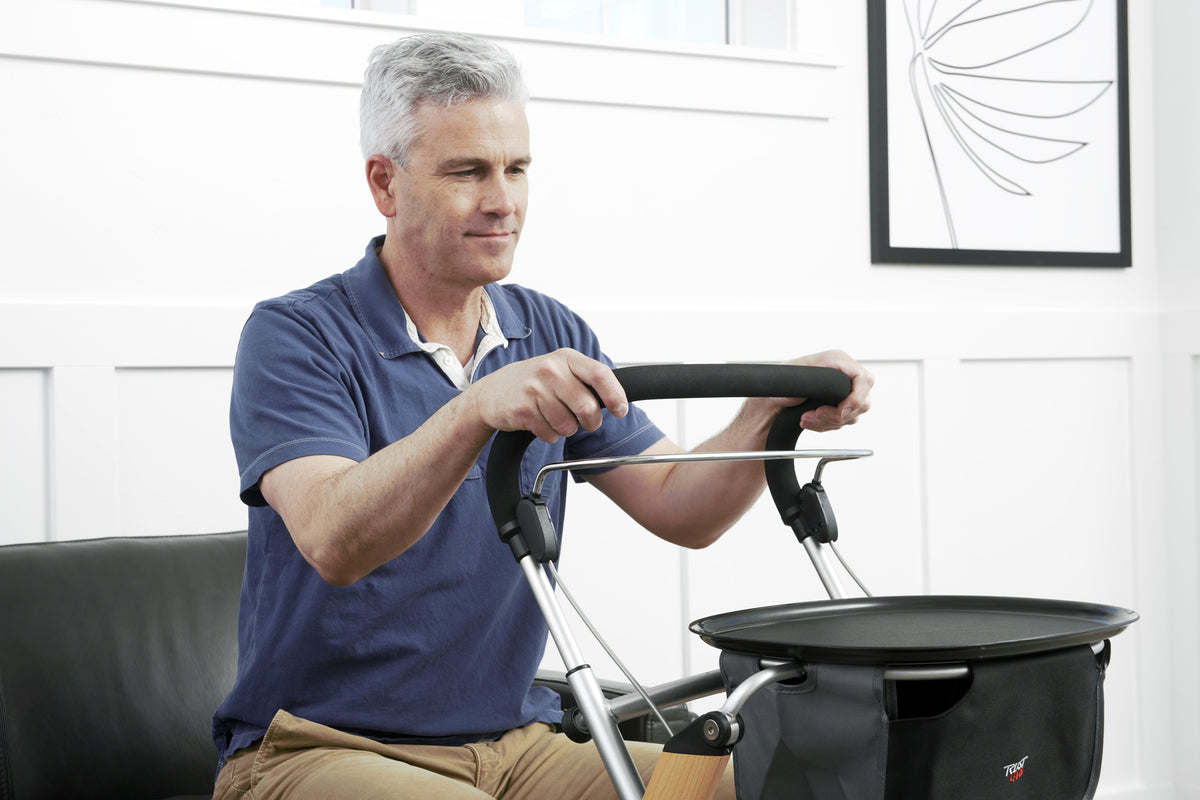 Indoor rollator with padded handles and attached tray bag, demonstrated by a seated man adjusting the handlebar indoors