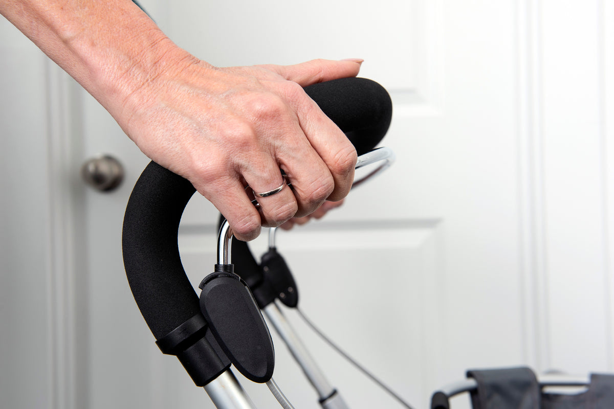 Rollator handle grip shown close up with a hand grasping the black foam padded handlebar indoors