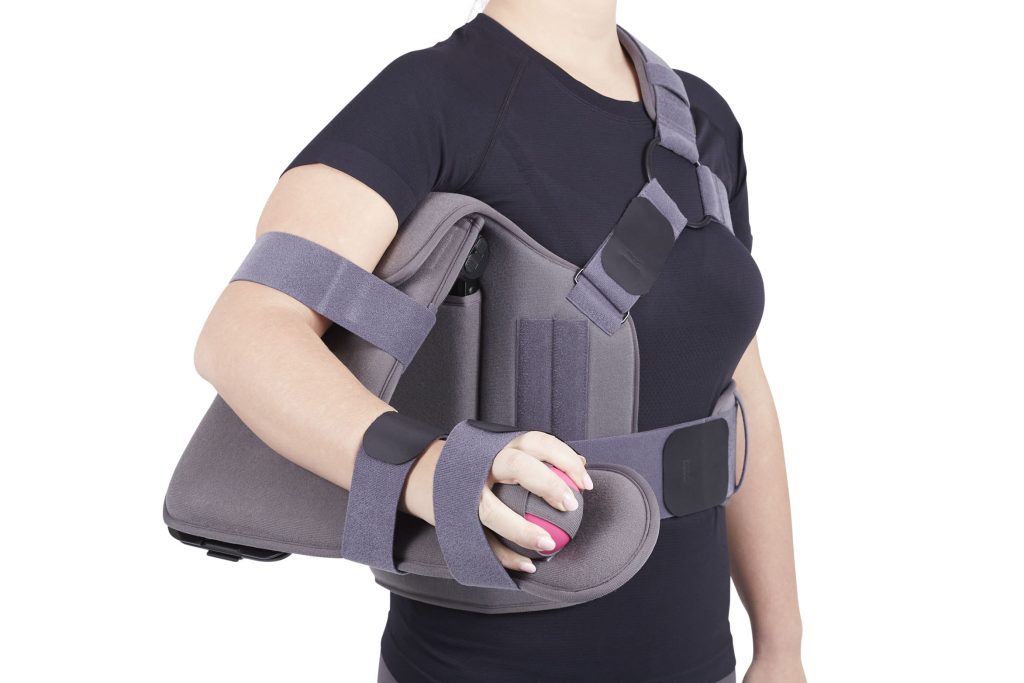 Shoulder ROM immobilizer worn by a woman in a black shirt, gray padded brace with chest straps and arm support cradle