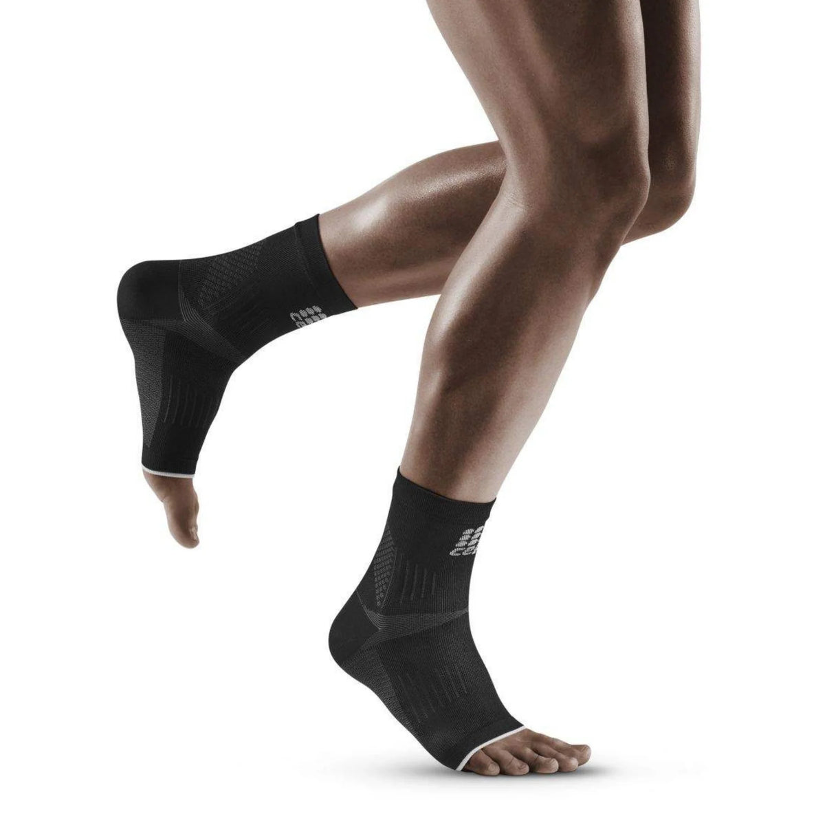 Ankle compression sleeves in black with grey accents worn on both feet of a person mid-stride, open-toe and open-heel design