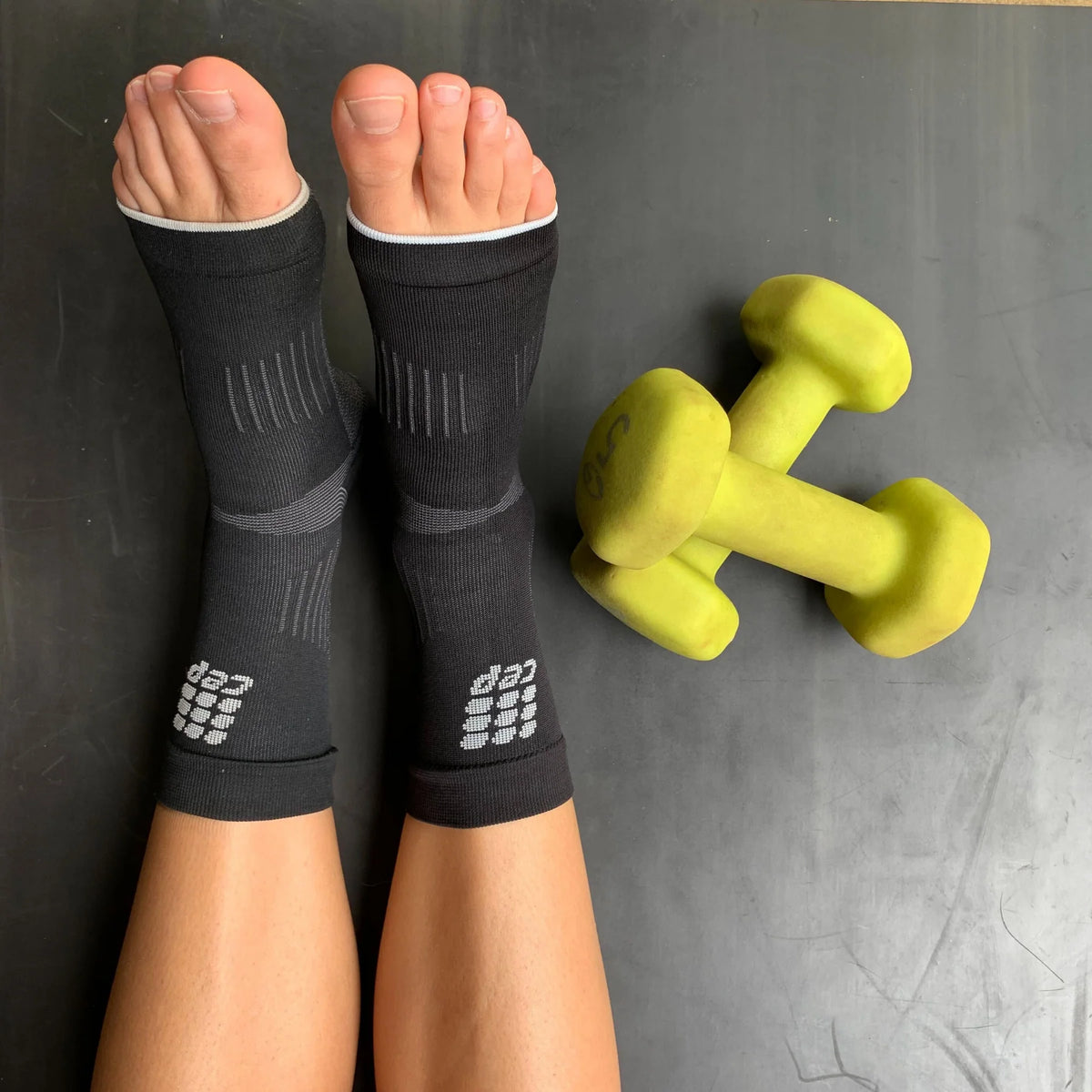 CEP compression ankle sleeves in black worn on both feet, shown from above beside two yellow dumbbells