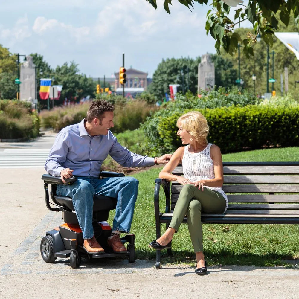 Pride Go Chair power wheelchair in orange and black, used by a man conversing with a woman on a park bench outdoors