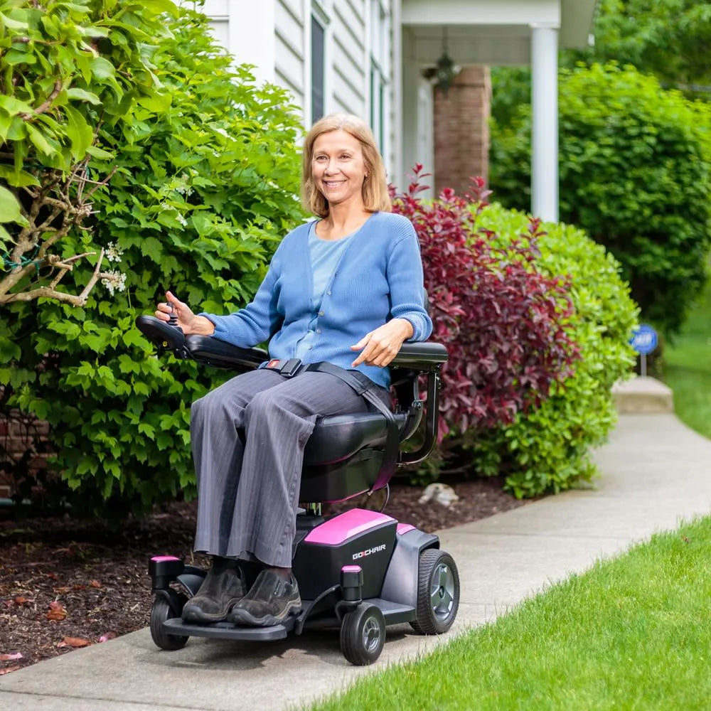 Power wheelchair in pink and black with joystick control, ridden outdoors on a sidewalk by a smiling woman near garden shrubs