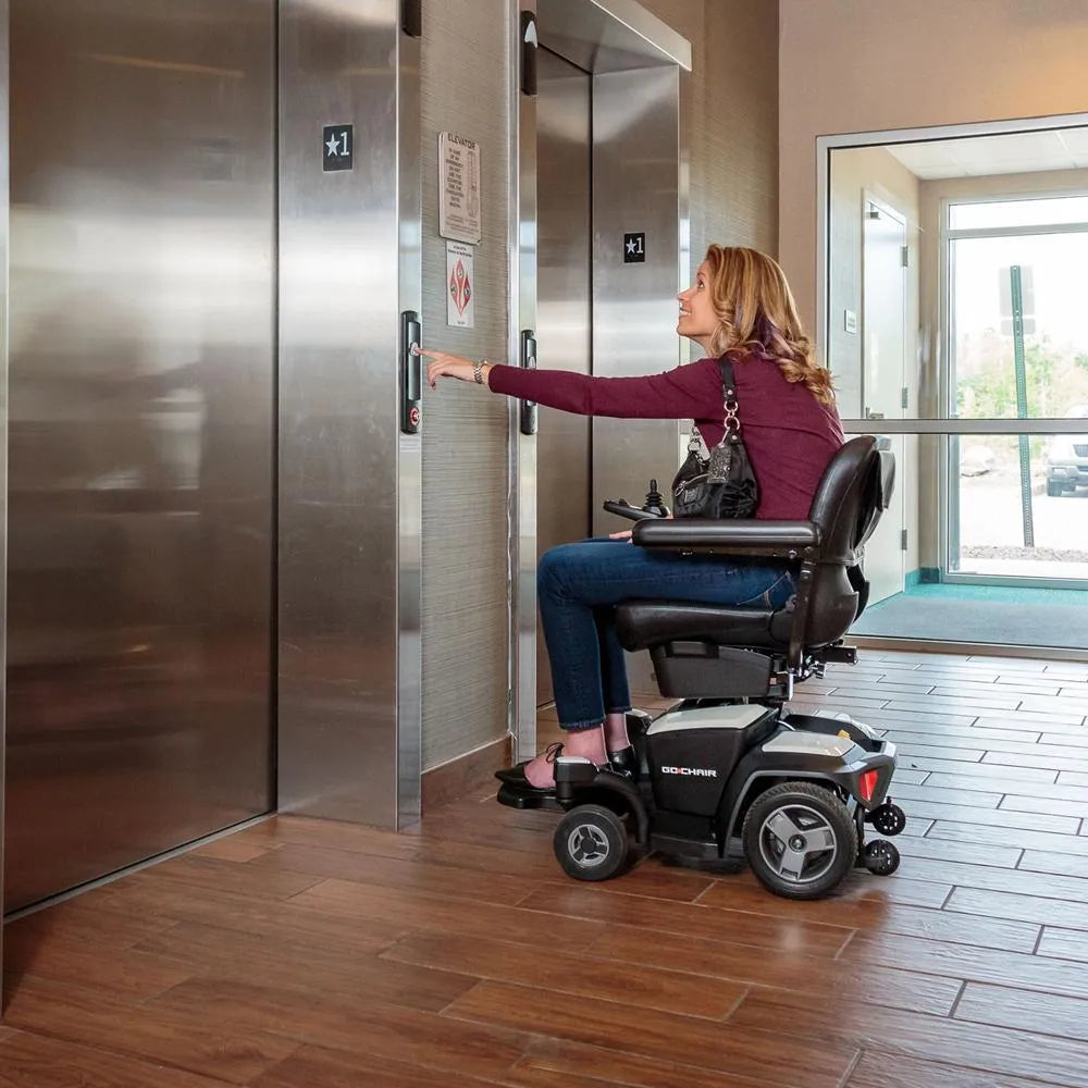 Power wheelchair with woman rider pressing elevator button in a building lobby, shown from the side