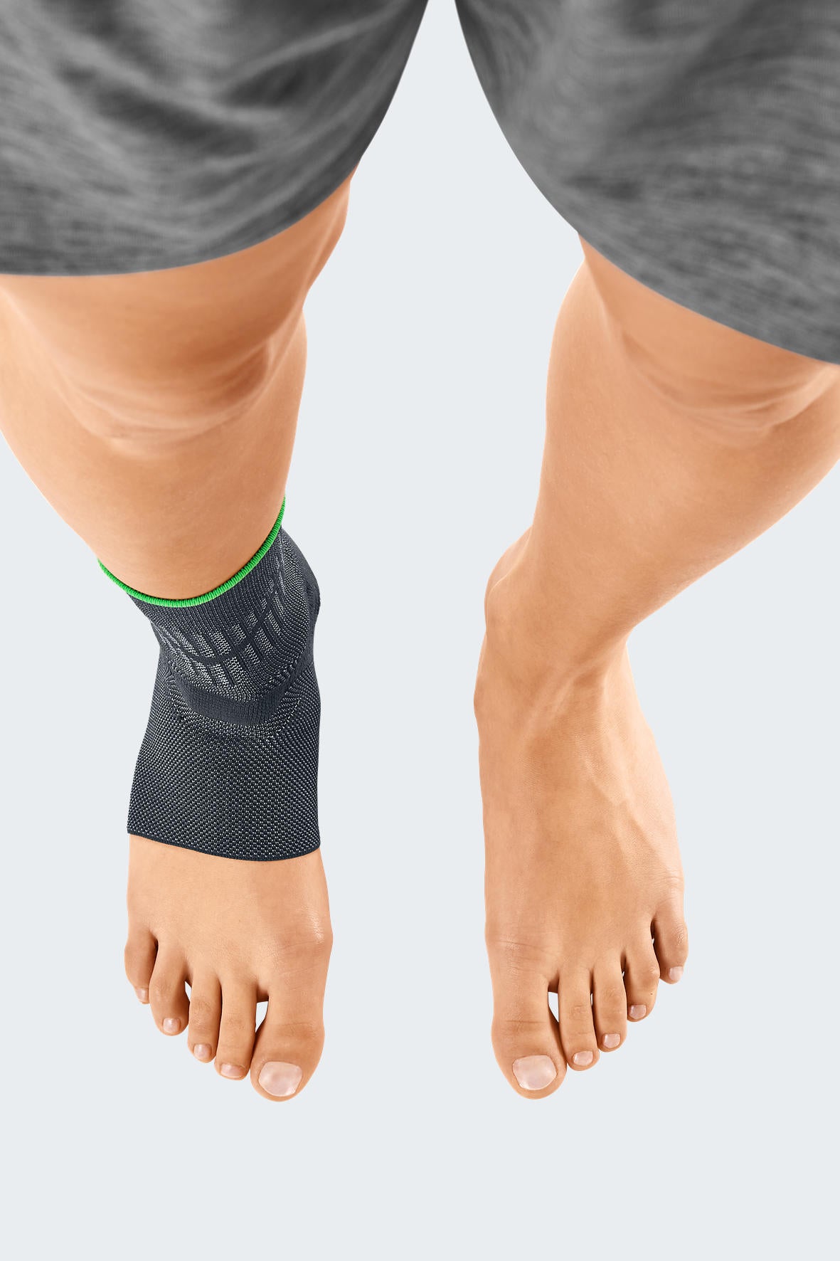 Ankle support sleeve in dark gray knit fabric with green trim, worn on left ankle, shown from above on both bare feet
