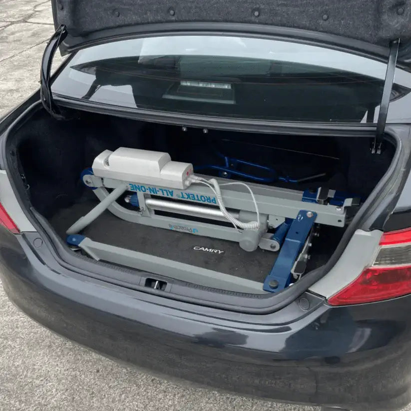 Patient car transfer lift folded and stored inside the open trunk of a sedan, demonstrating compact portability