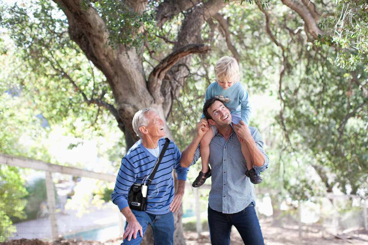 Portable oxygen concentrator worn via shoulder strap by older man walking outdoors with adult and child among trees