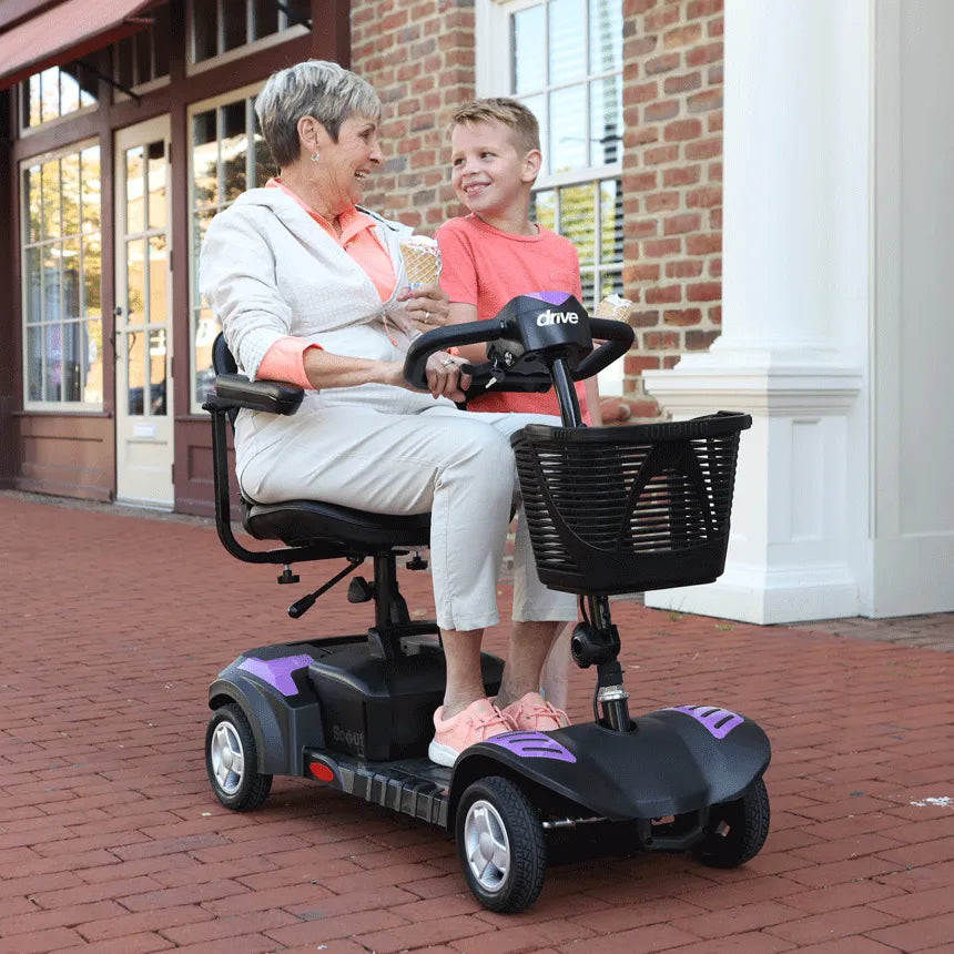 Power mobility scooter with black and purple accents, older woman riding outdoors on brick sidewalk beside a smiling child