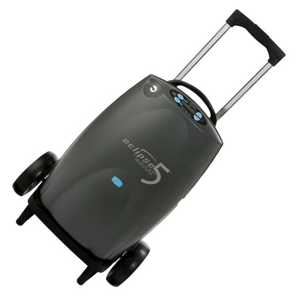 Portable oxygen concentrator in dark gray with rolling wheels, retractable handle, carry strap, and blue control buttons on
