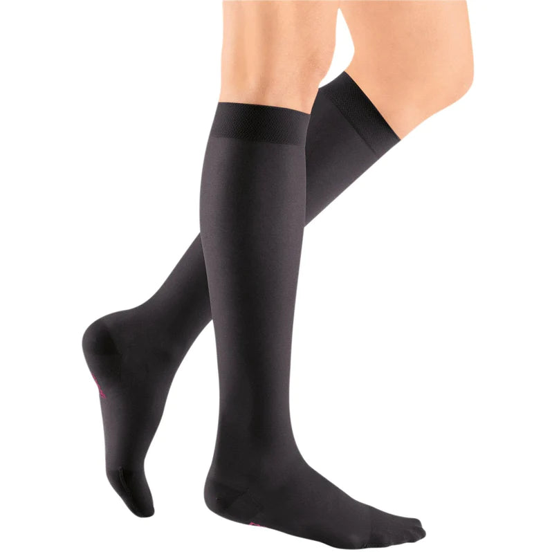 Compression stockings in black knee-high closed-toe style with wide comfort band top, shown on a person's legs against white