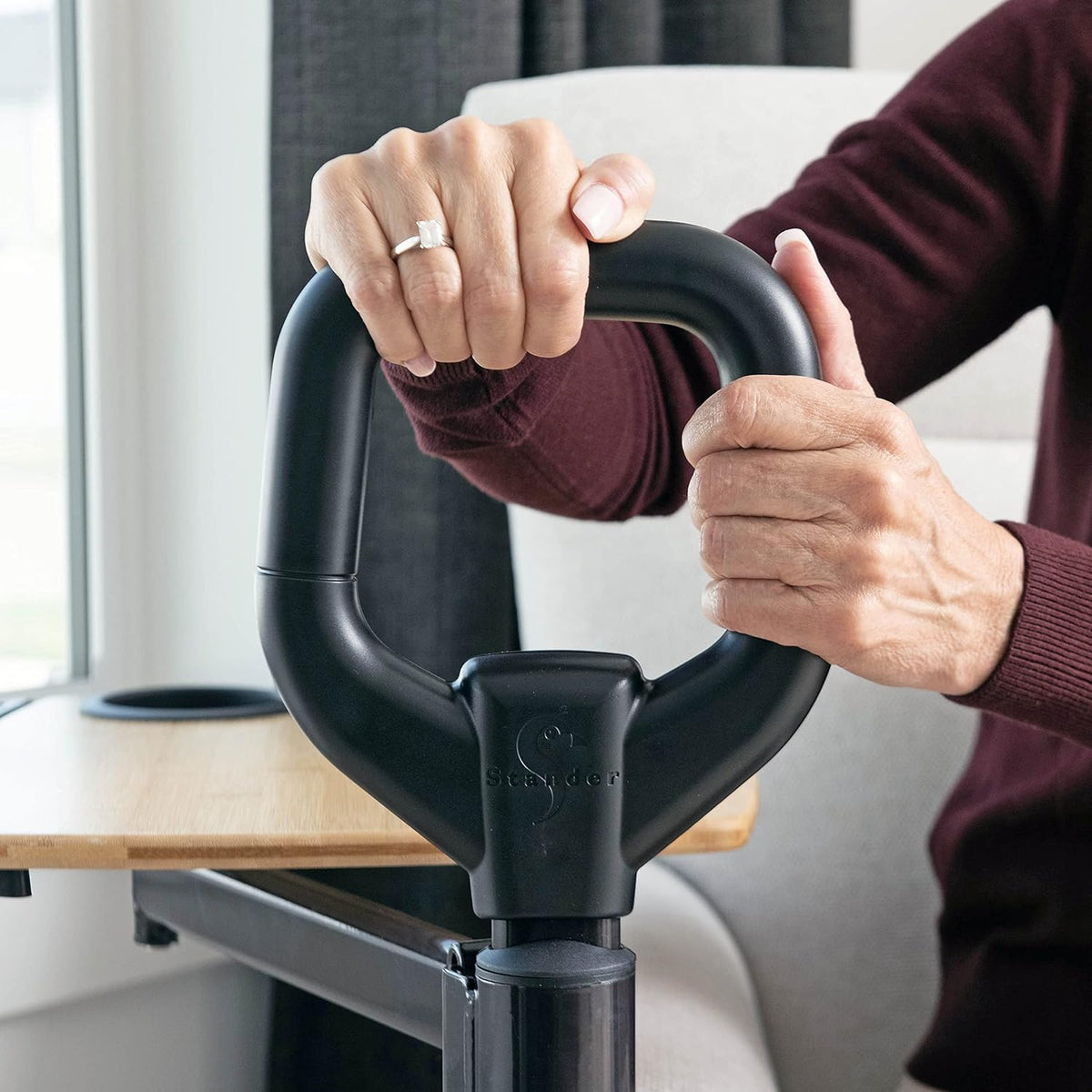 Stander Assist-A-Tray stand assist handle with loop grip shown being gripped by both hands near a couch and overbed table