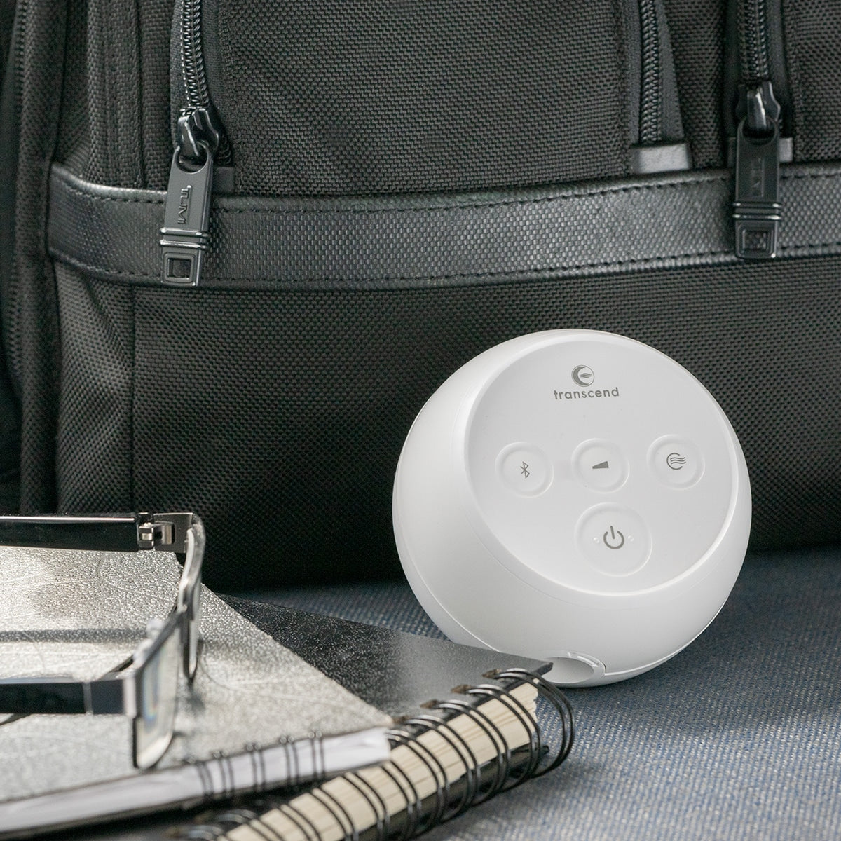 Transcend Micro Auto CPAP machine, small white round device with control buttons, shown beside a travel bag and notebook