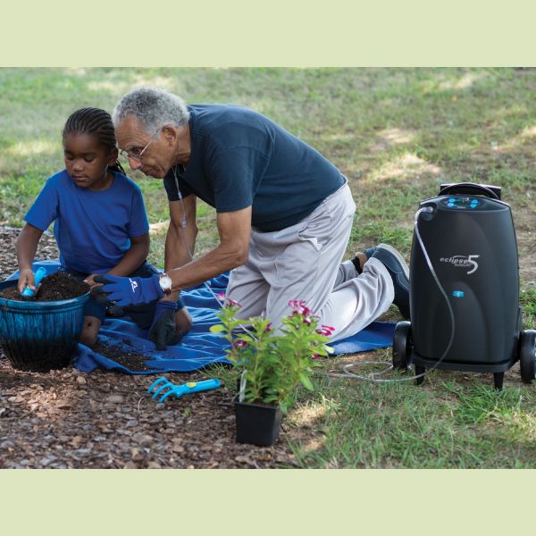 Eclipse 5 portable oxygen concentrator on wheels outdoors beside an elderly man and child gardening together