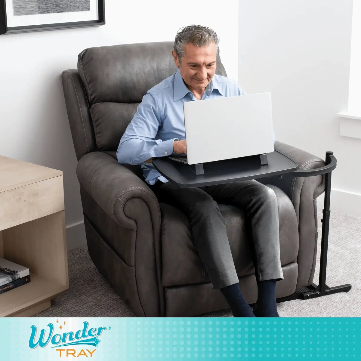 Overbed tray table with black surface beside a recliner as man uses a laptop while seated in a gray leather chair