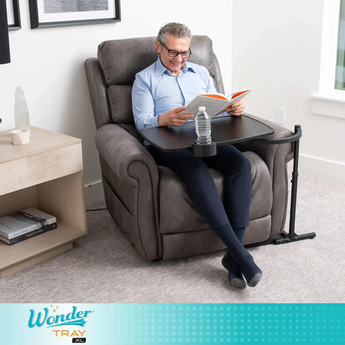 Overbed tray table with cup holder positioned over recliner armrest, used by seated man reading a book with water bottle on