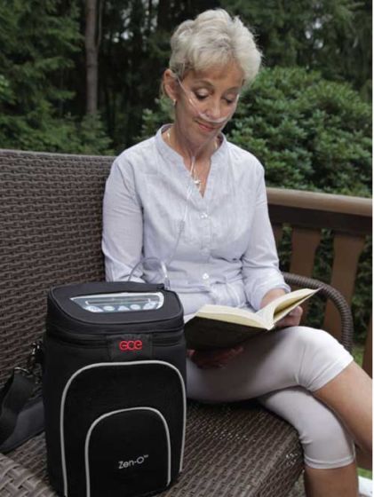 Zen-O portable oxygen concentrator in black carrying case beside a woman wearing a nasal cannula reading on an outdoor patio