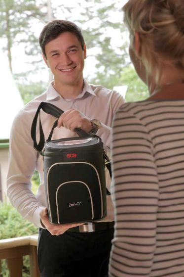 Zen-O portable oxygen concentrator in black carrying case with shoulder strap, held by smiling man at doorway