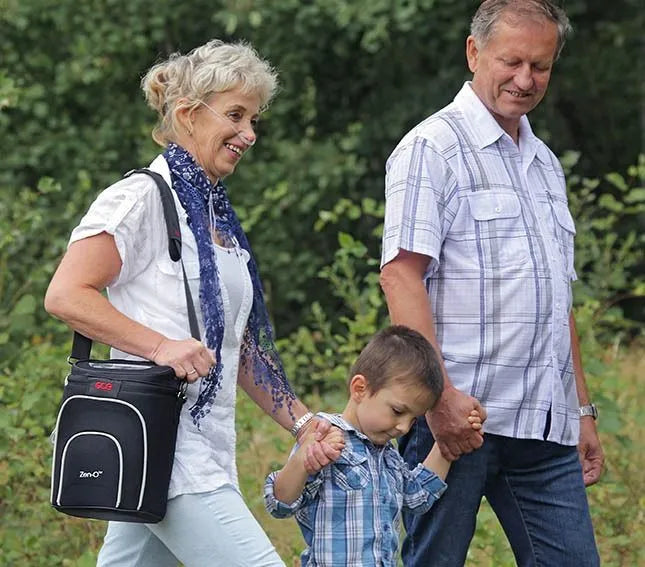 Portable oxygen concentrator carried in a black shoulder bag by a smiling woman walking outdoors with a man and young child