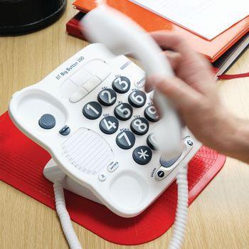 Non-slip pad in red positioned under a large-button telephone on a wooden desk, shown with a hand lifting the handset