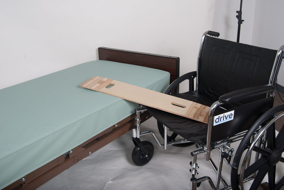 Bariatric transfer board bridging a hospital bed and Drive wheelchair, showing cut-out hand grips on a wide wooden plank